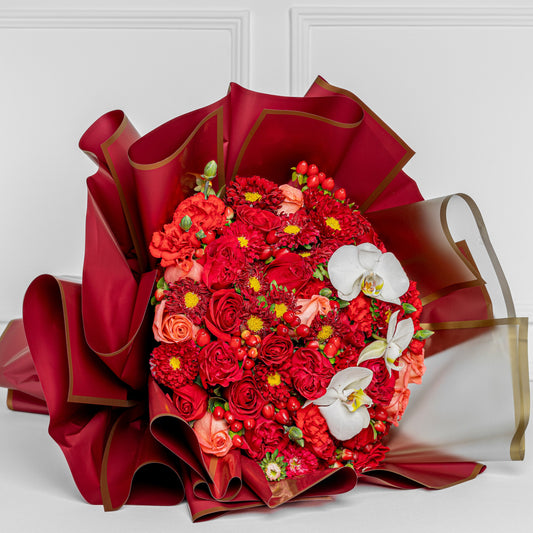 Elegant bouquet with red roses, coral flowers, and white orchids wrapped in burgundy paper with gold details – Medellín same-day flower delivery.