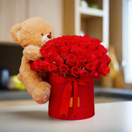 Arrangement of premium red roses in a decorative box with a red ribbon, accompanied by a soft teddy bear, perfect for Valentine’s Day in Medellín.