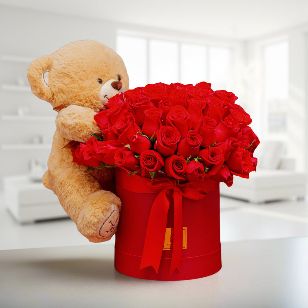 Arrangement of premium red roses in a decorative box with a red ribbon, accompanied by a soft teddy bear, perfect for Valentine’s Day in Medellín.