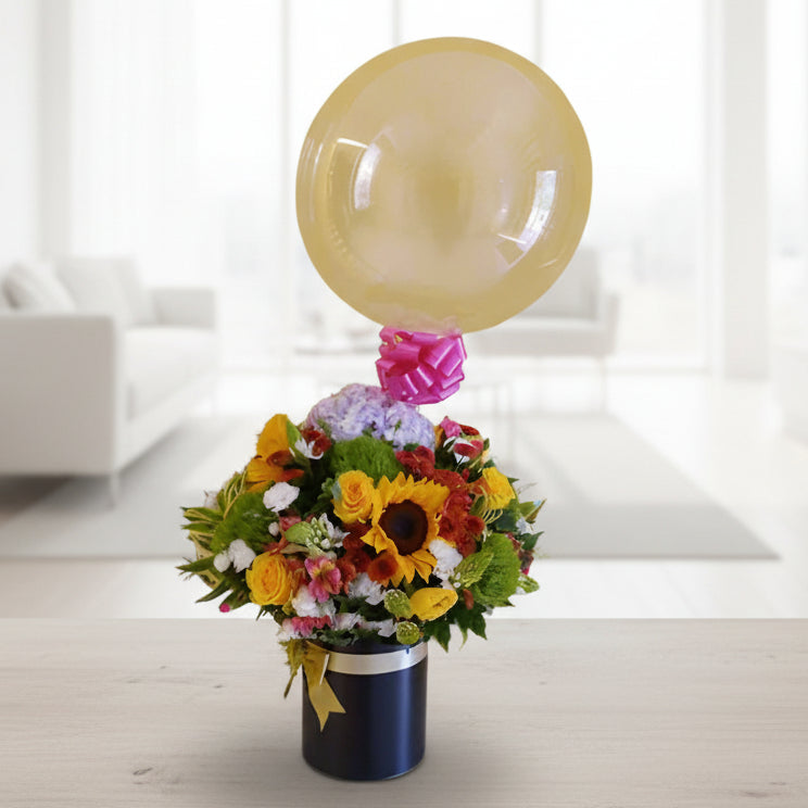 Charming birthday floral arrangement with multicolor roses, seasonal flowers, and a transparent 'Feliz Cumpleaños' balloon in a pink hatbox. Perfect for delivery in Cali.