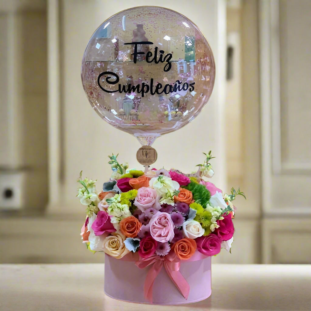 Charming birthday floral arrangement with multicolor roses, seasonal flowers, and a transparent 'Feliz Cumpleaños' balloon in a pink hatbox. Perfect for delivery in Cali.