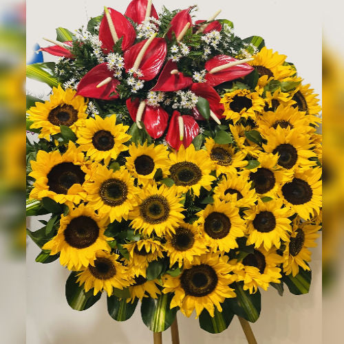 Funeral wreath with sunflowers, red anthuriums, and seasonal foliage on a wooden tripod, includes a customizable funeral ribbon, available for delivery in Medellín, Colombia.
