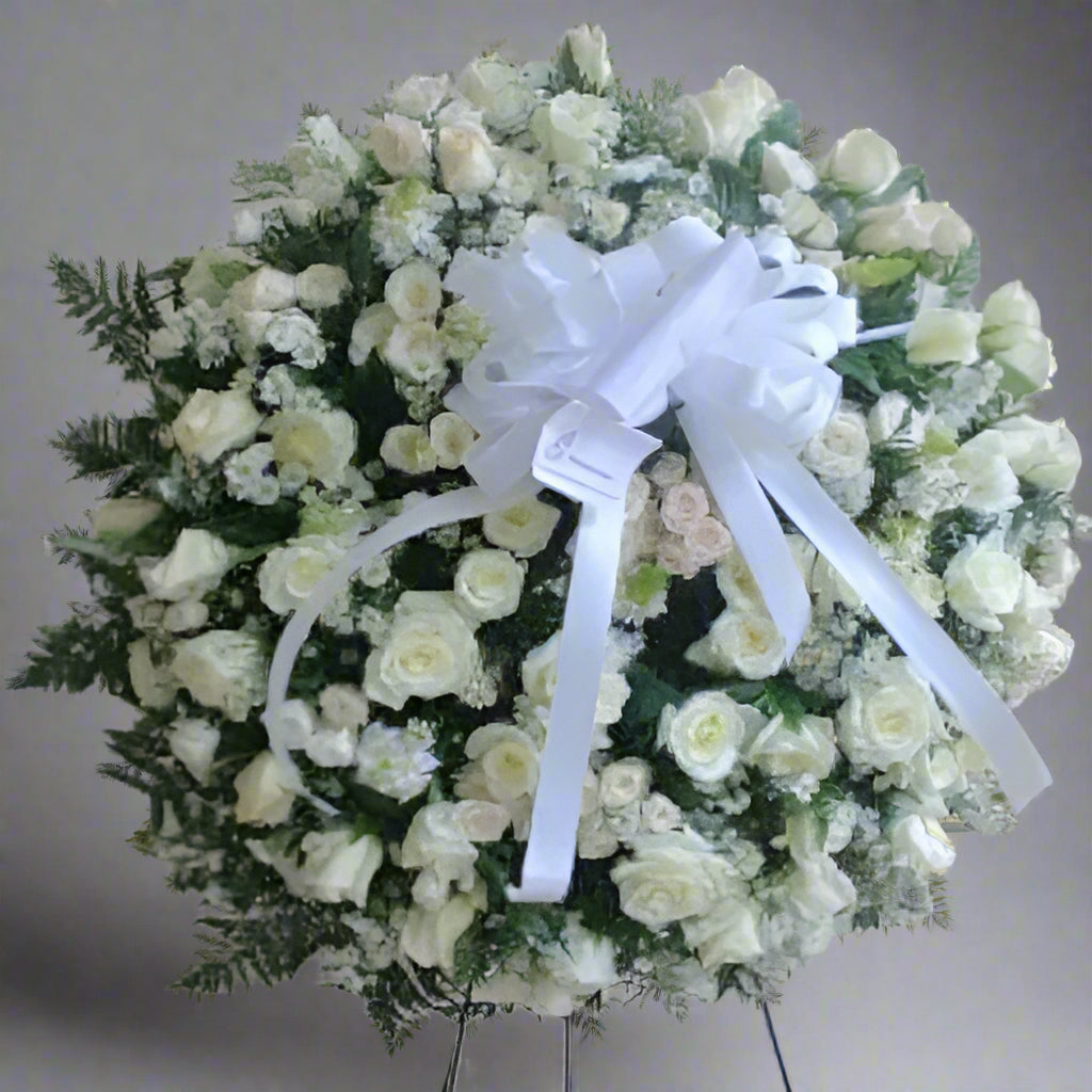 Elegant standing funeral wreath made of white roses, greenery, and a large white ribbon – available for delivery across Colombia.