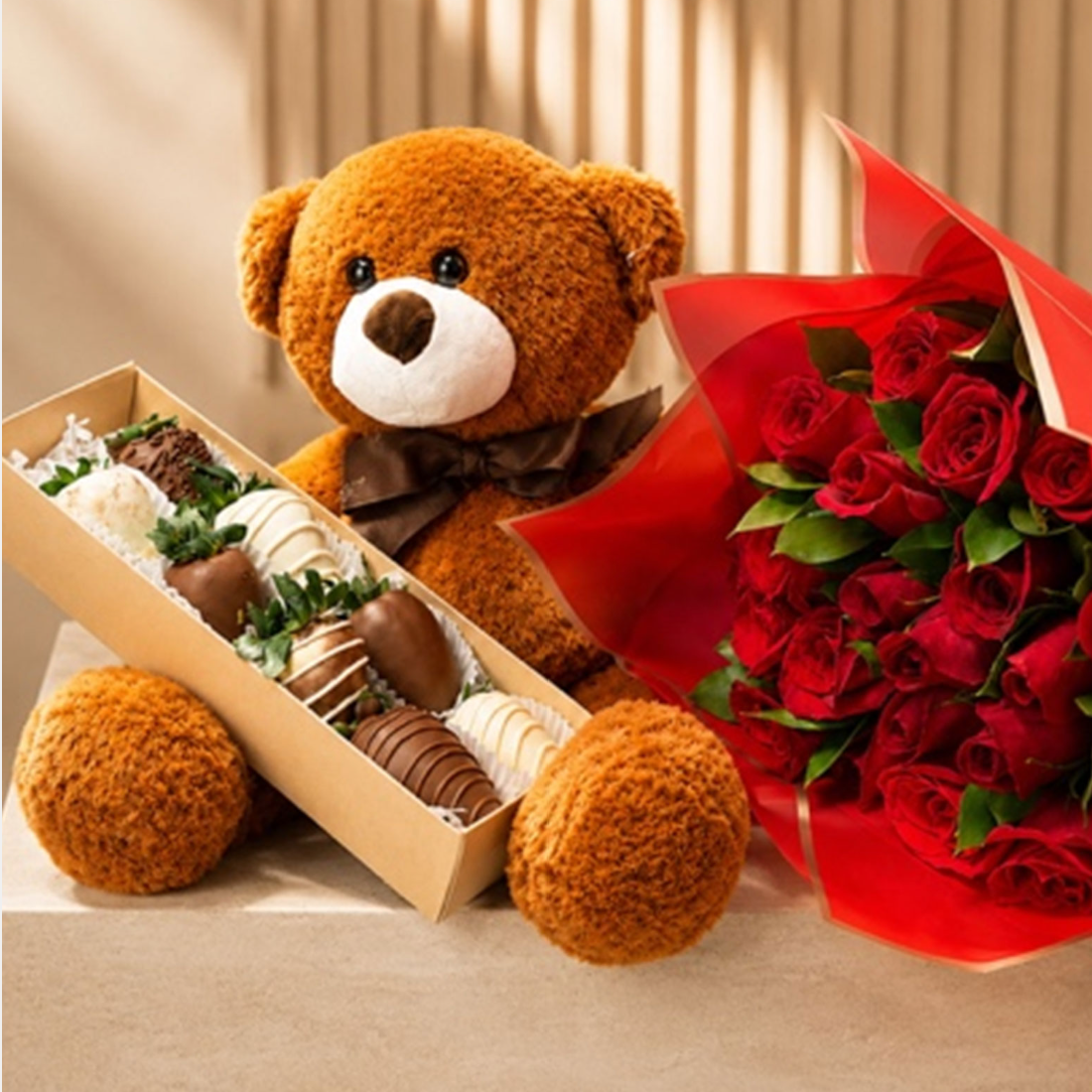 A brown plush teddy bear with a white snout and dark brown bow tie sits beside a long box of assorted chocolates and a large bouquet of red roses wrapped in red paper Medellin